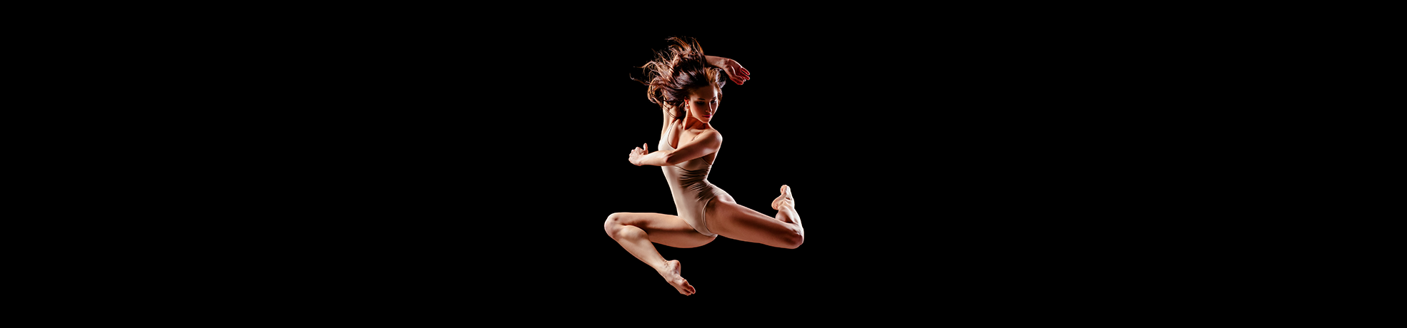 Dancewear collection: A dancer in a beige leotard leaps high against a black background.