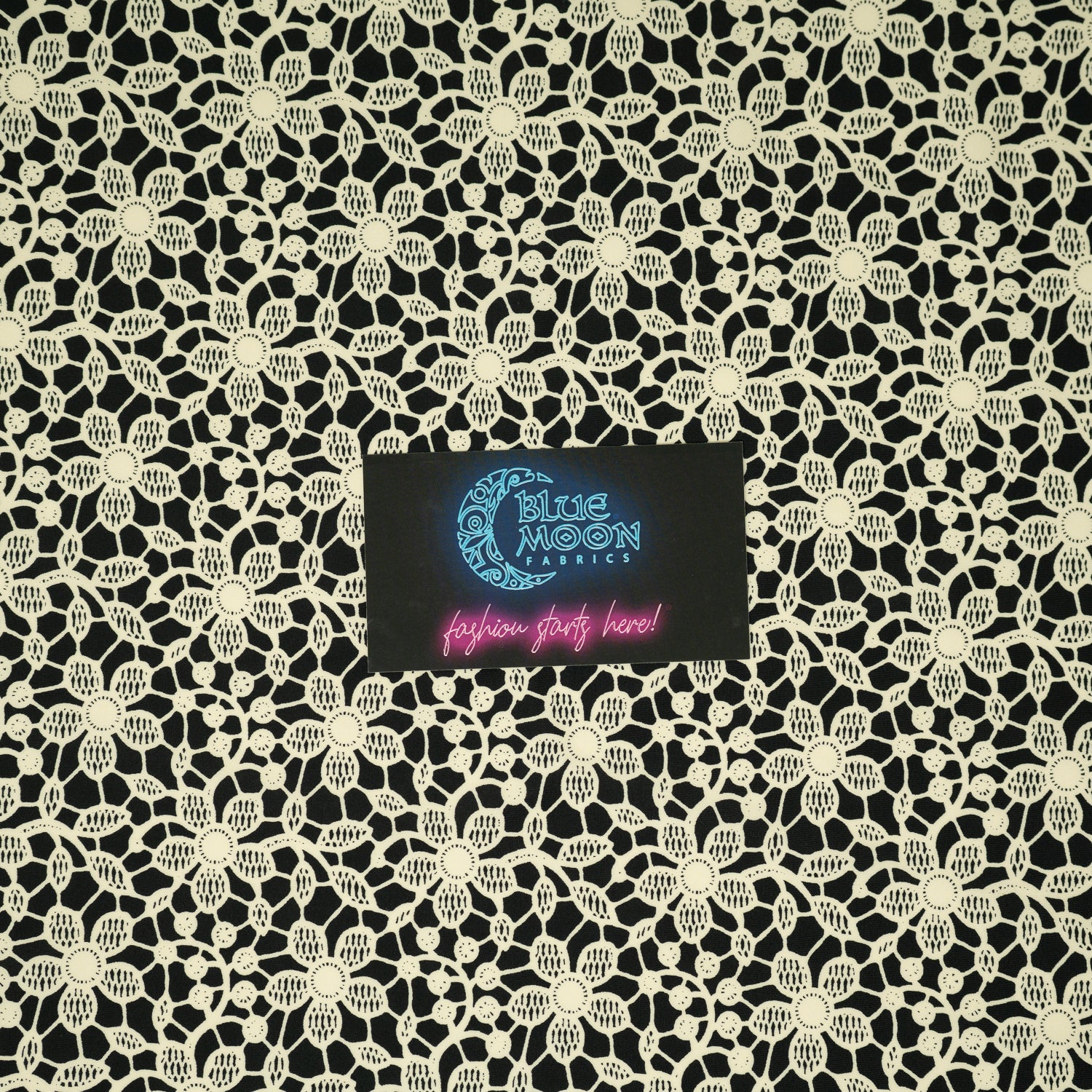 A flat sample of Ivory Flower Lace Pattern on Black Printed Spandex with a Blue Moon Fabrics standard size business card laid on top of the print for pattern sizing.