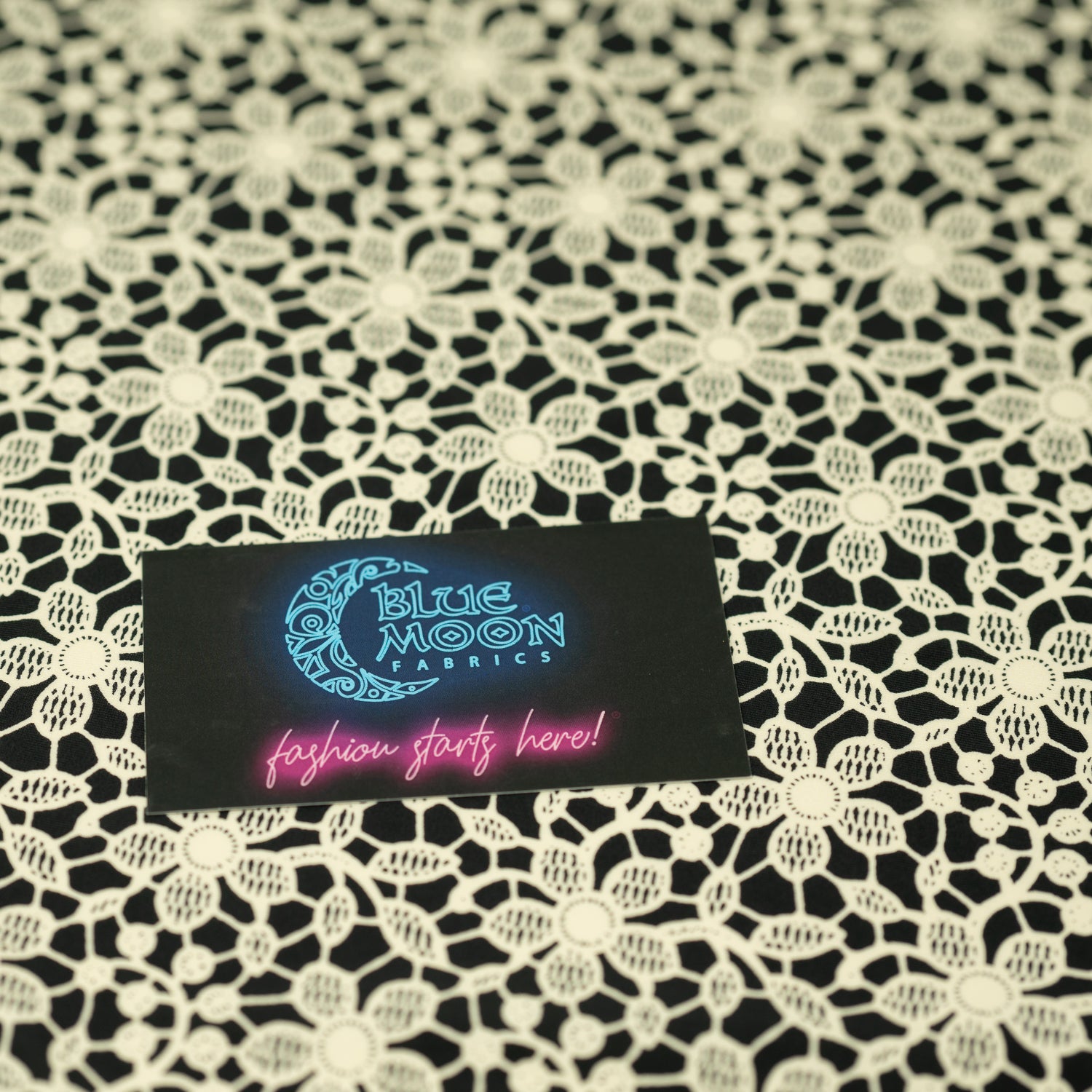 A flat sample of Ivory Flower Lace Pattern on Black Printed Spandex with a Blue Moon Fabrics standard size business card laid on top of the print for pattern sizing.