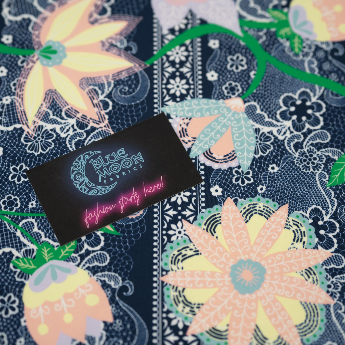 A flat sample of Chantilly Floral Lace Printed Spandex with a Blue Moon Fabrics standard size business card laid on top of the print for pattern sizing.